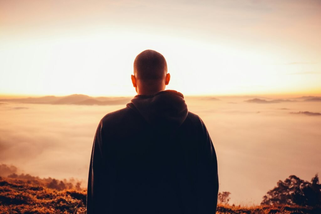 A solitary man in a hoodie stands gazing at a serene sunrise above a sea of clouds.